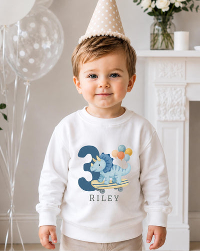 A young boy wearing a white sweatshirt with a dinosaur and balloons on it, standing in front of a fireplace with balloons and a vase of flowers.