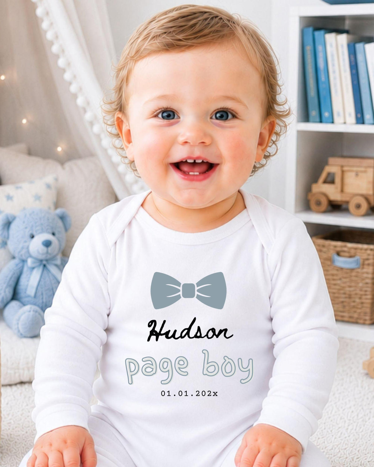 A smiling baby boy wearing a white onesie with a bow tie and the name "Hudson" printed on it, sitting in a room with a teddy bear and bookshelf in the background.