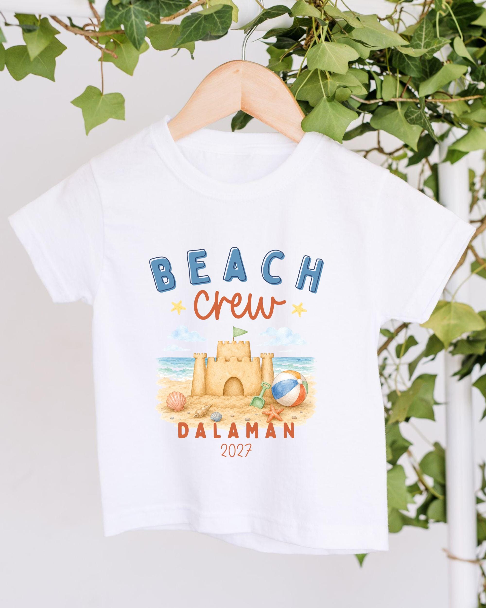 A white t-shirt with a beach-themed design hanging on a wooden hanger, with a sand castle and beach ball depicted.