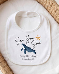 A white bib with a graphic of a whale and a starfish, along with the text "Sea You Soon" and "Baby Davidson December 2026".