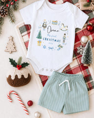 A white onesie with a Christmas-themed design, a pair of green shorts, and various Christmas decorations such as a pine cone, a small Christmas tree, and ornaments are arranged on a plaid blanket.