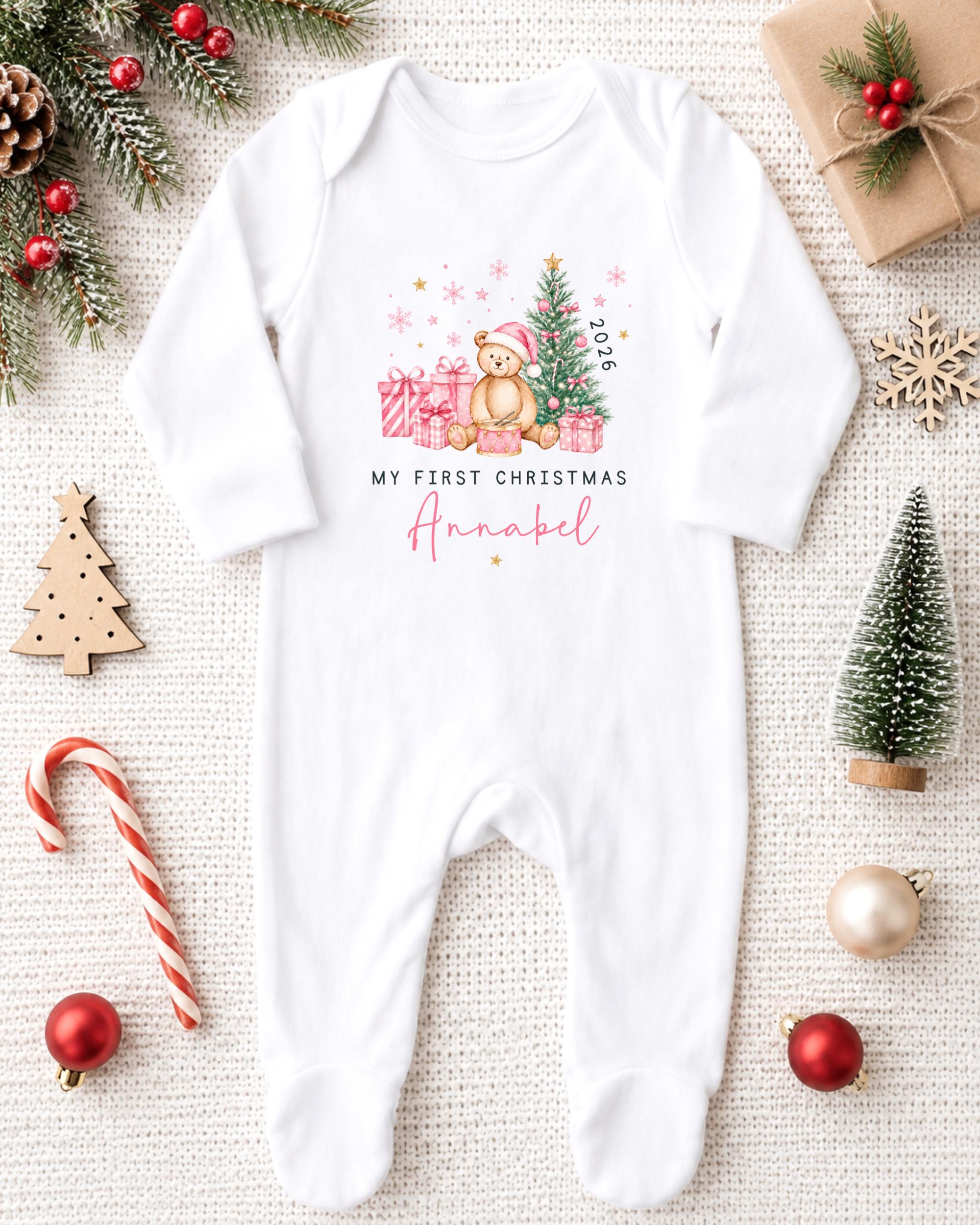 A white onesie with a teddy bear and Christmas decorations, including a small Christmas tree, presents, and ornaments, arranged on a white background.