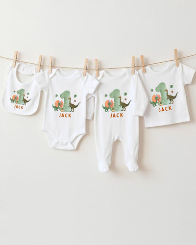 A collection of white baby clothes, including bodysuits and pajamas, with dinosaur designs and the name "Jack" printed on them.