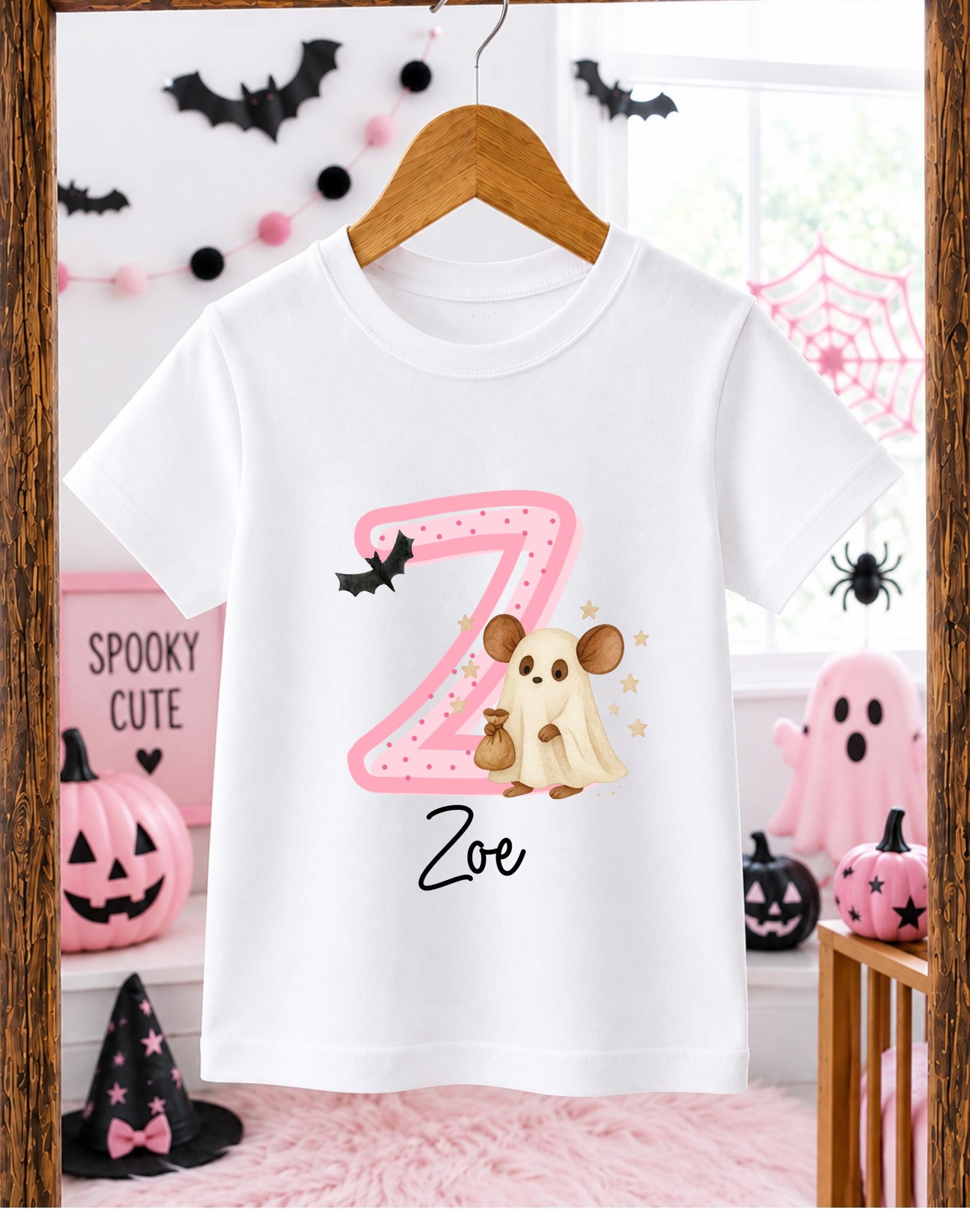 A white t-shirt with a pink and black design featuring a ghost and a bat, along with the name "Zoe" printed on it. The t-shirt is hanging on a wooden hanger, and there are Halloween decorations and pumpkins visible in the background.