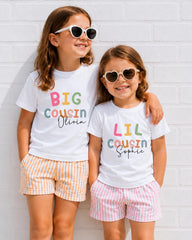 Two young girls wearing white t-shirts with the text "BIG COUSIN Olivia" and "LIL COUSIN Sophie" printed on them, smiling and posing together against a white brick wall.