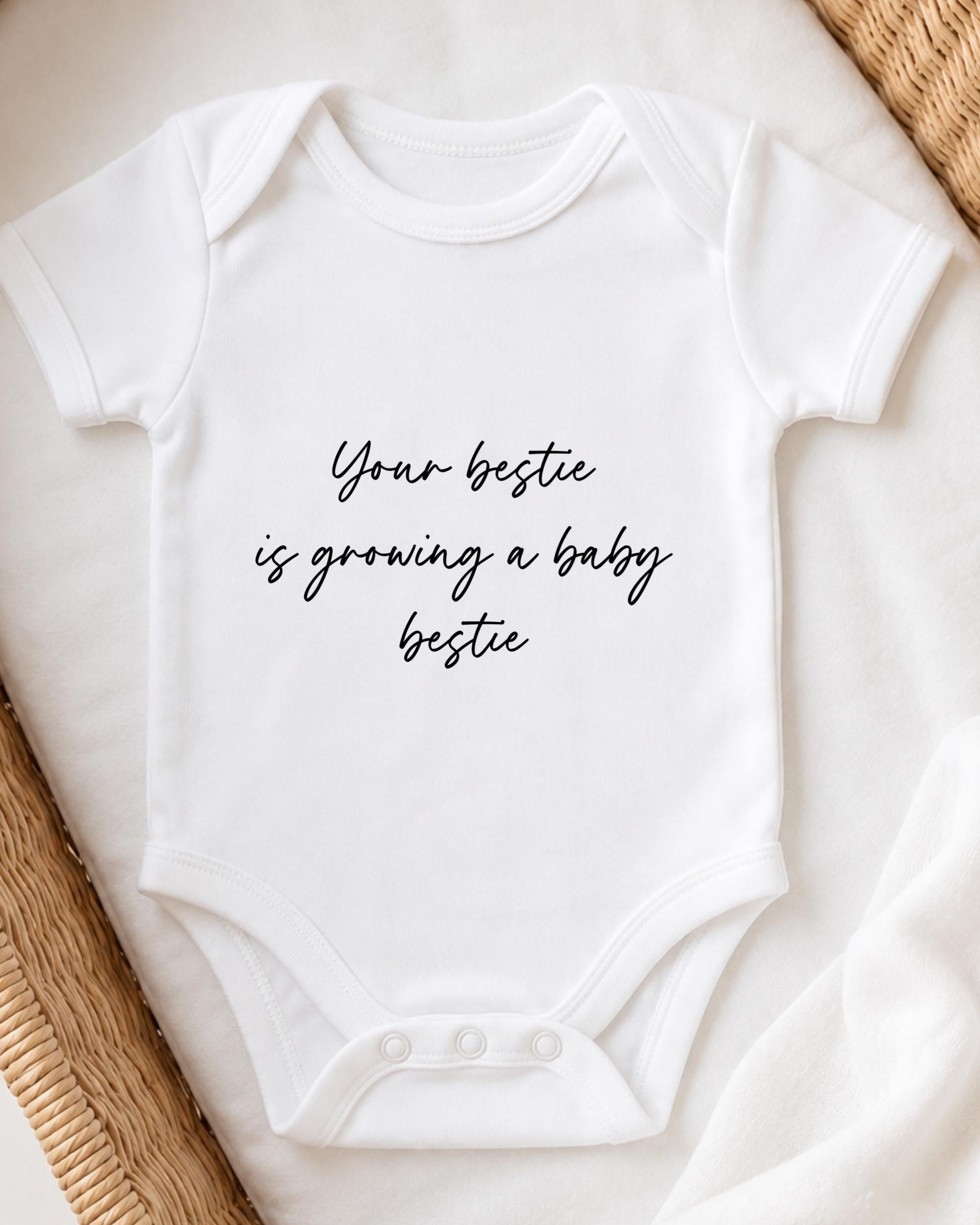 A white baby onesie with the text "Your bestie is growing a baby bestie" printed on it.