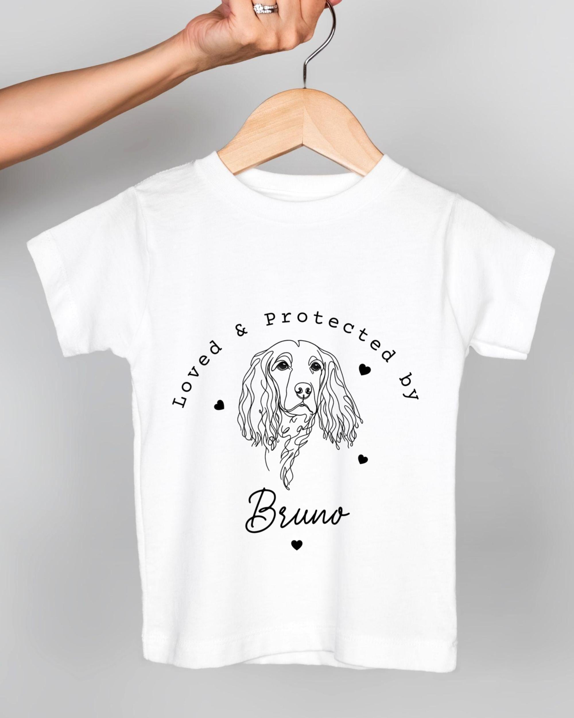 A white t-shirt with a graphic design of a dog's head and the text "Loved & protected by Bruno" printed on it.