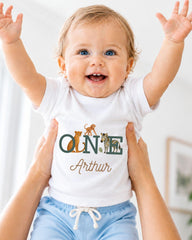 A smiling baby wearing a white t-shirt with the name "Arthur" and animal illustrations, being held up by someone's arms.