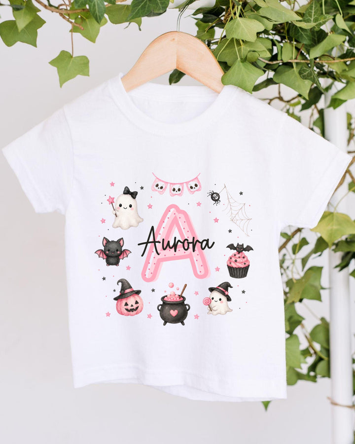 A white t-shirt with a personalized design featuring a pink "A" surrounded by various Halloween-themed illustrations, including a ghost, bats, a spider, a pumpkin, a witch's hat, a pumpkin, a cupcake, and a spider web.