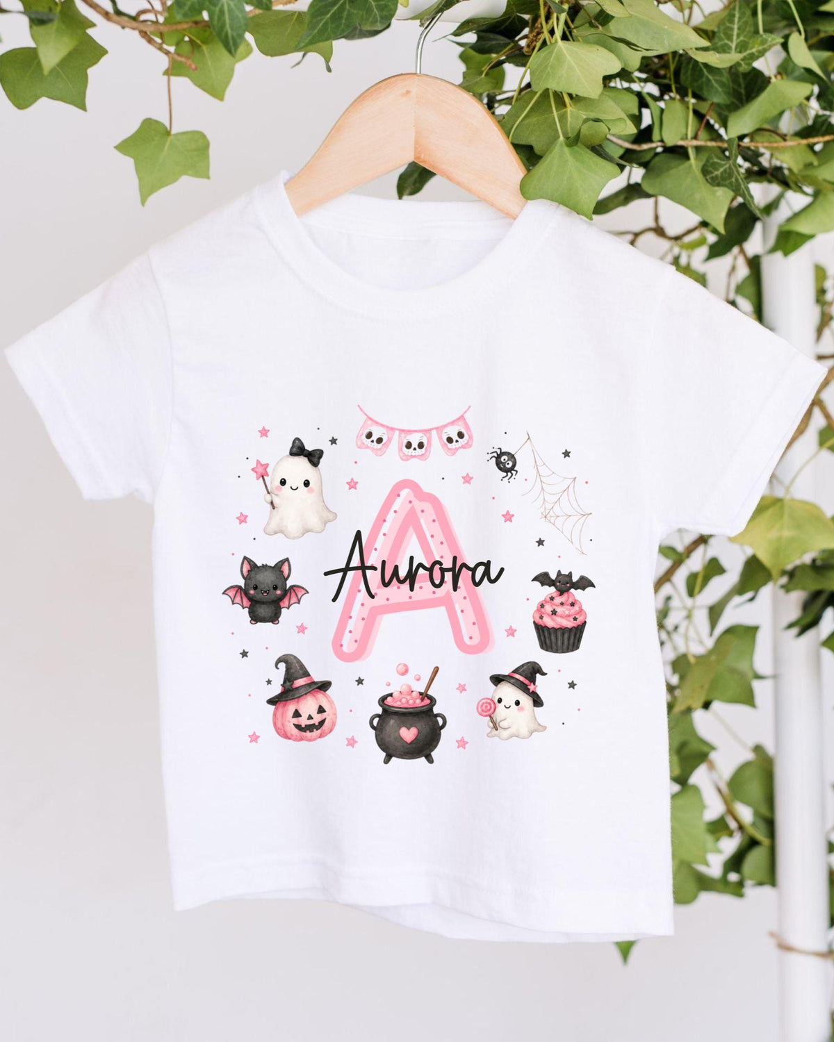 A white t-shirt with a personalized design featuring a pink "A" surrounded by various Halloween-themed illustrations, including a ghost, bats, a spider, a pumpkin, a witch's hat, a pumpkin, a cupcake, and a spider web.