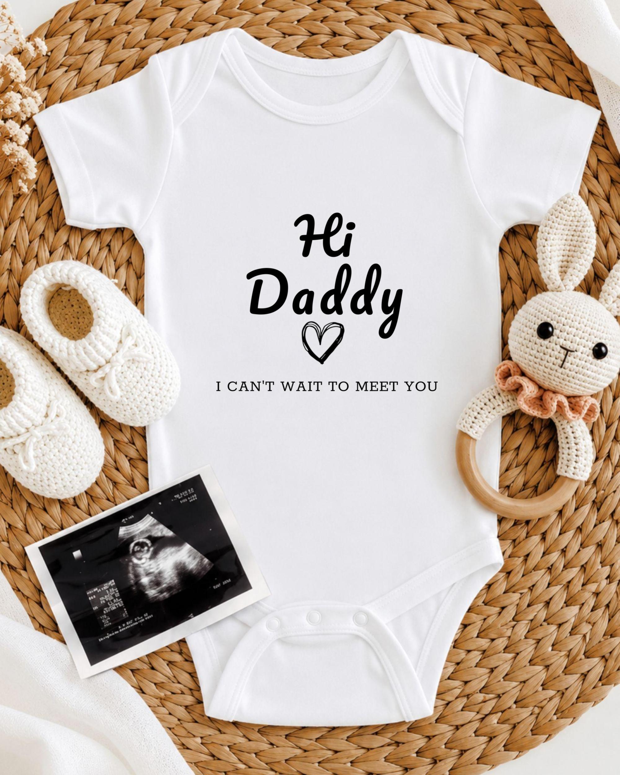 A baby onesie with the text "Hi Daddy" and a heart symbol, along with a crocheted bunny toy and a baby bootie, all arranged on a woven placemat.