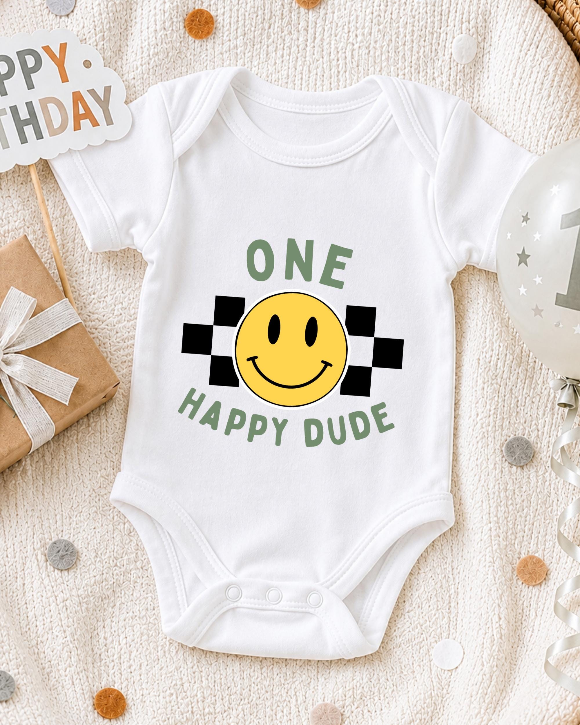 A white onesie with a yellow smiley face and the text "ONE HAPPY DUDE" printed on it, along with a "Happy Birthday" sign and a birthday balloon.