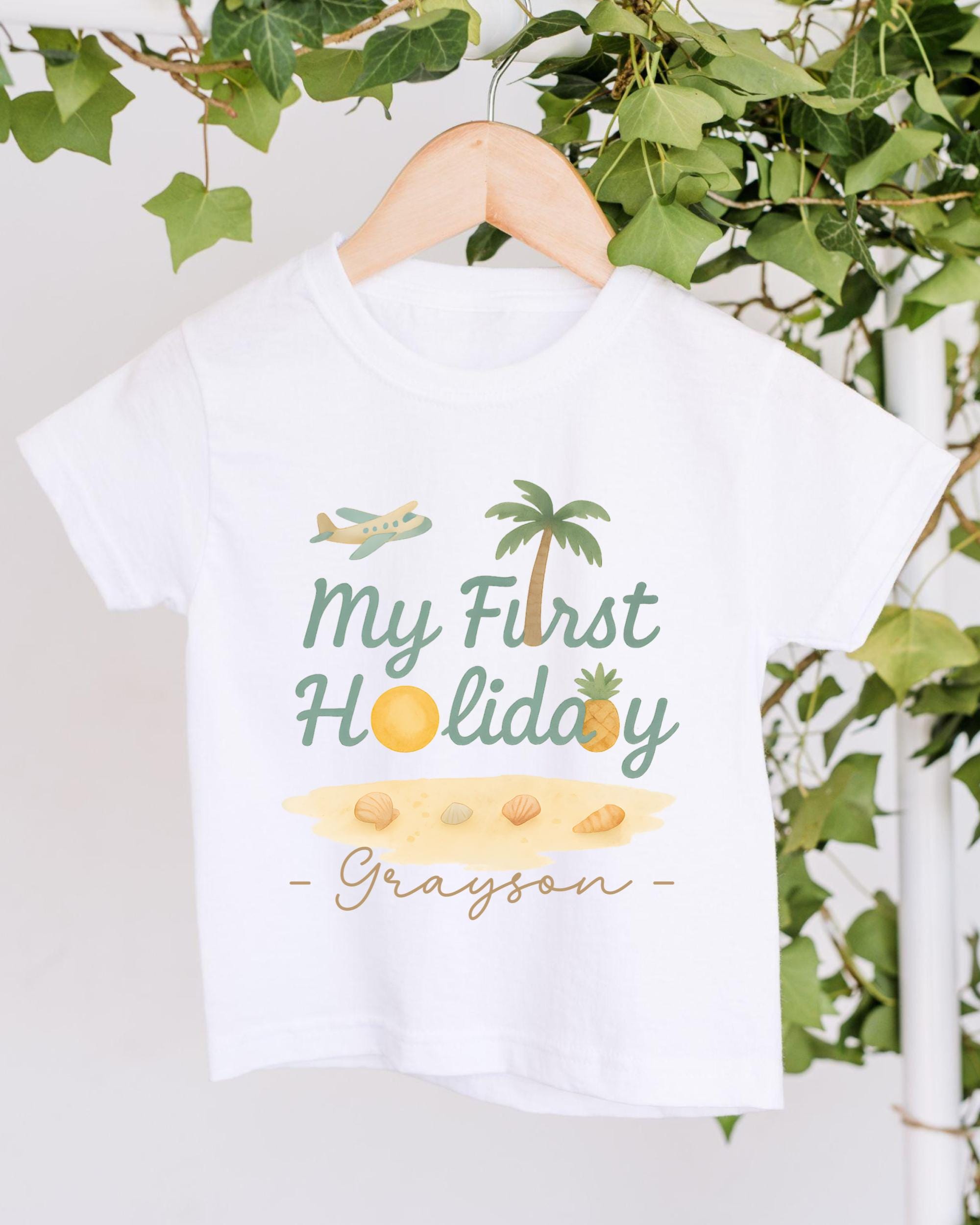 A white t-shirt with a graphic design featuring a palm tree, a plane, and various beach-related items, along with the text "My First Holiday - Grayson".