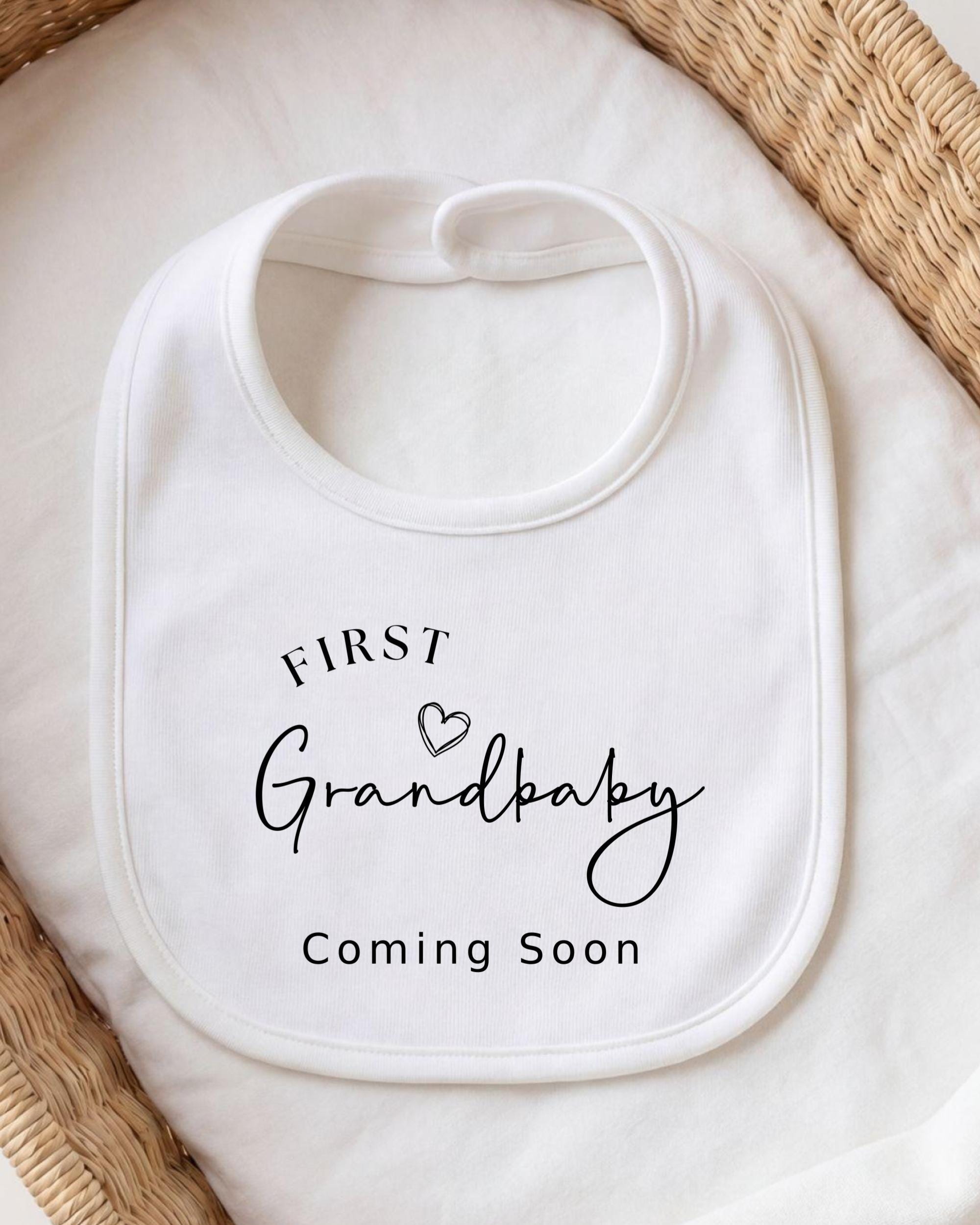 A white bib with the text "FIRST Grandbaby Coming Soon" printed on it, placed on a white cloth.