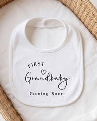 A white bib with the text "FIRST Grandbaby Coming Soon" printed on it, placed on a white cloth.