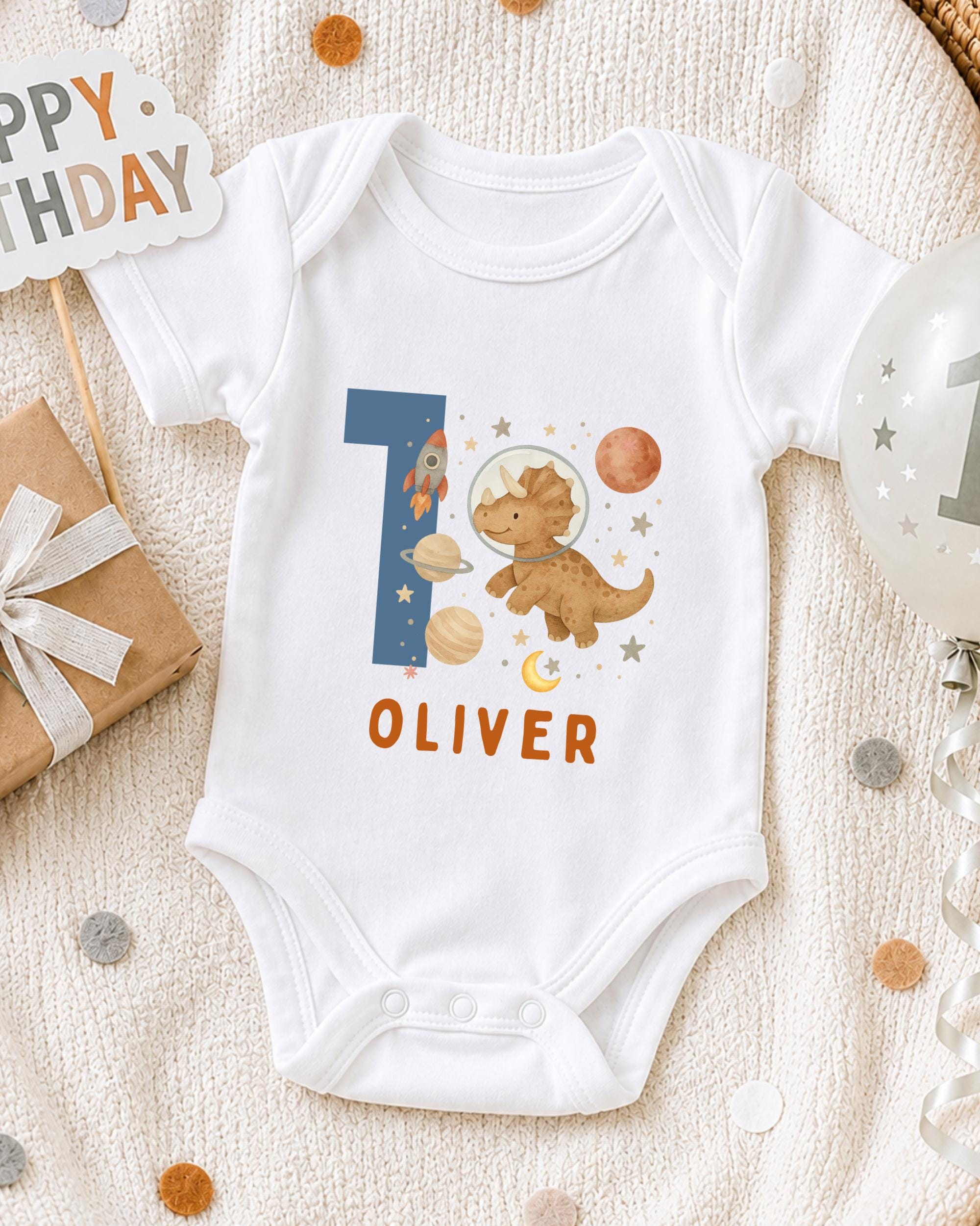 A white onesie with a dinosaur and space-themed design, along with a "Happy Birthday Oliver" sign, is displayed on a blanket.