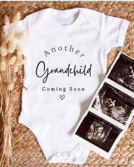 A white baby onesie with the text "Another Grandchild Coming Soon" printed on it, along with a heart symbol. Next to the onesie is a set of three black and white baby ultrasound images.