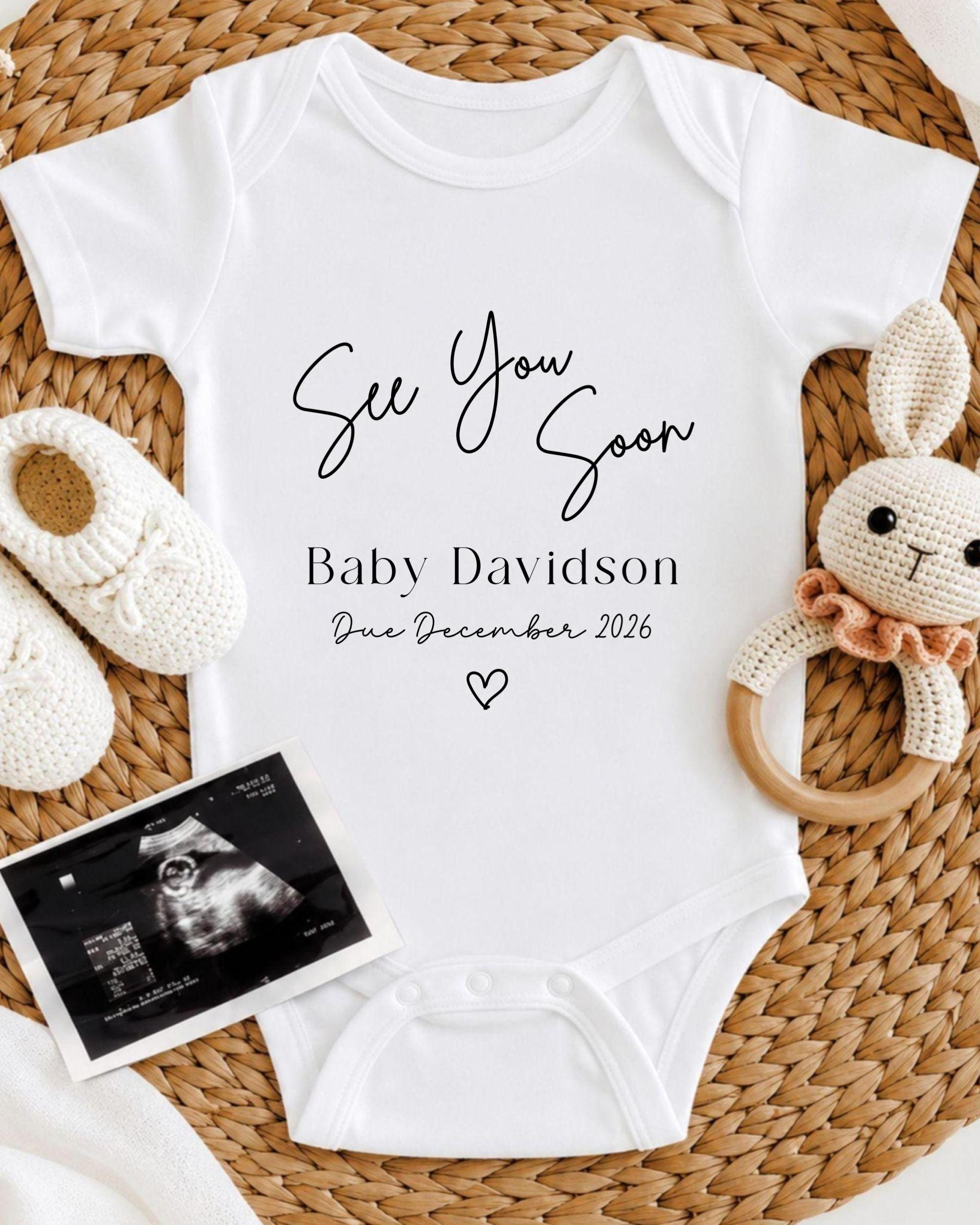a white baby onesie with the text "See You Soon Baby Davidson Due December 2026" printed on it, along with a crocheted bunny toy and a black and white photograph of an infant's head.
