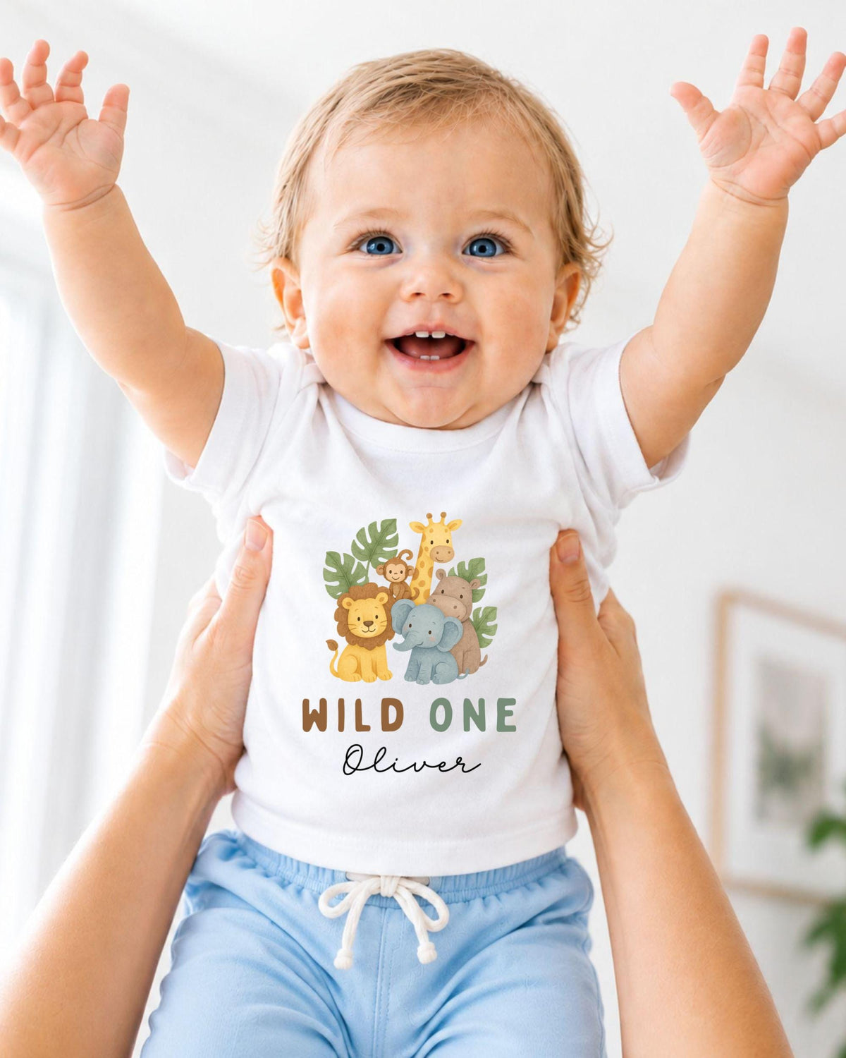 A smiling baby wearing a white t-shirt with a jungle-themed design and the text "WILD ONE Oliver" is being held up by someone's arms.