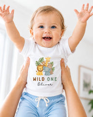 A smiling baby wearing a white t-shirt with a jungle-themed design and the text "WILD ONE Oliver" is being held up by someone's arms.