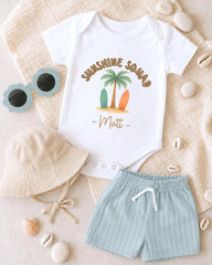 A baby's outfit, including a white onesie with a palm tree design and the name "Matt", a beige bucket hat, and light blue shorts, is laid out on a beige surface alongside sunglasses and seashells.