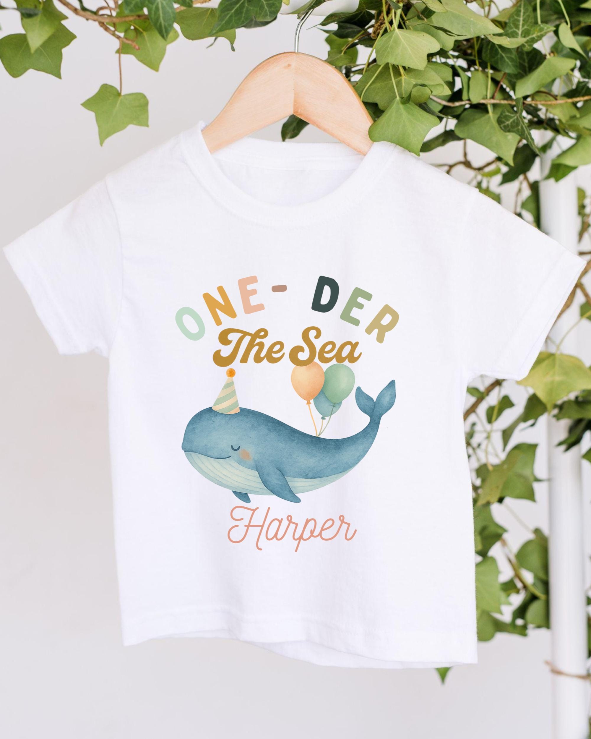 A white t-shirt with a colorful graphic design featuring a whale wearing a party hat and balloons, along with the text "One-DER The Sea Harper".