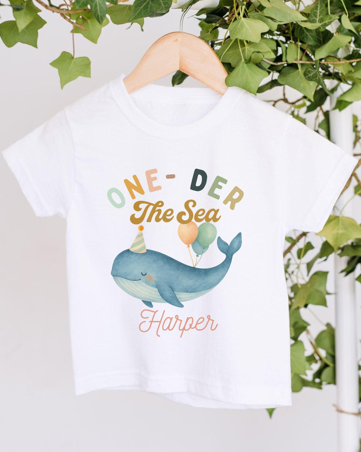 A white t-shirt with a colorful graphic design featuring a whale wearing a party hat and balloons, along with the text "One-DER The Sea Harper".