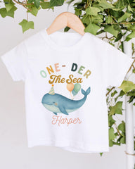 A white t-shirt with a colorful graphic design featuring a whale wearing a party hat and balloons, along with the text "One-DER The Sea Harper".