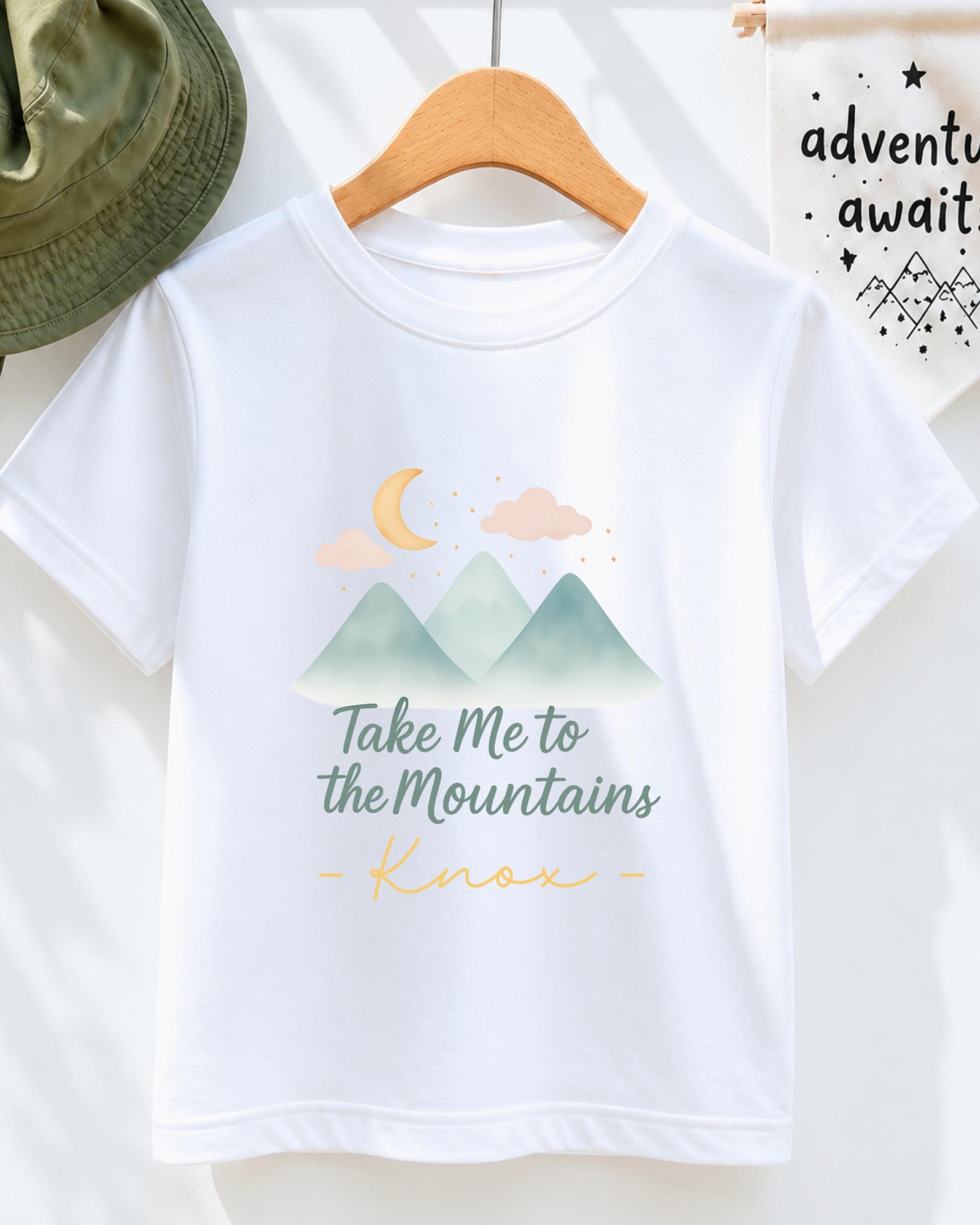 A white t-shirt with a graphic design of mountains and a crescent moon, along with the text "Take Me to the Mountains" and "Knox" printed on it.