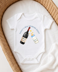 A white onesie with a graphic design depicting a bottle of wine and a bottle of milk, accompanied by the text "DADDY'S NEW DRINKING BUDDY".