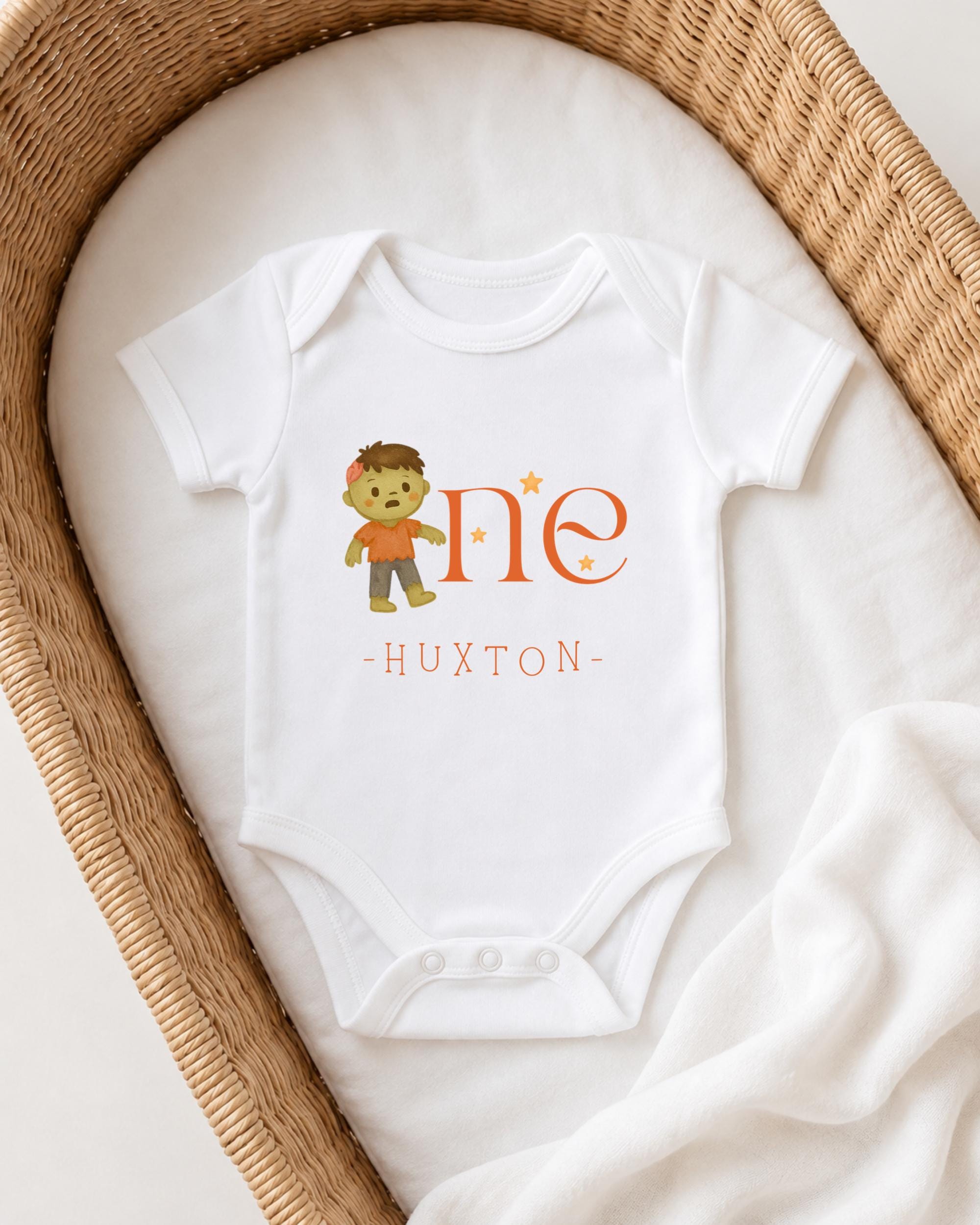 A white onesie with a cartoon character and the text "HUXTON" printed on it, placed in a woven basket.