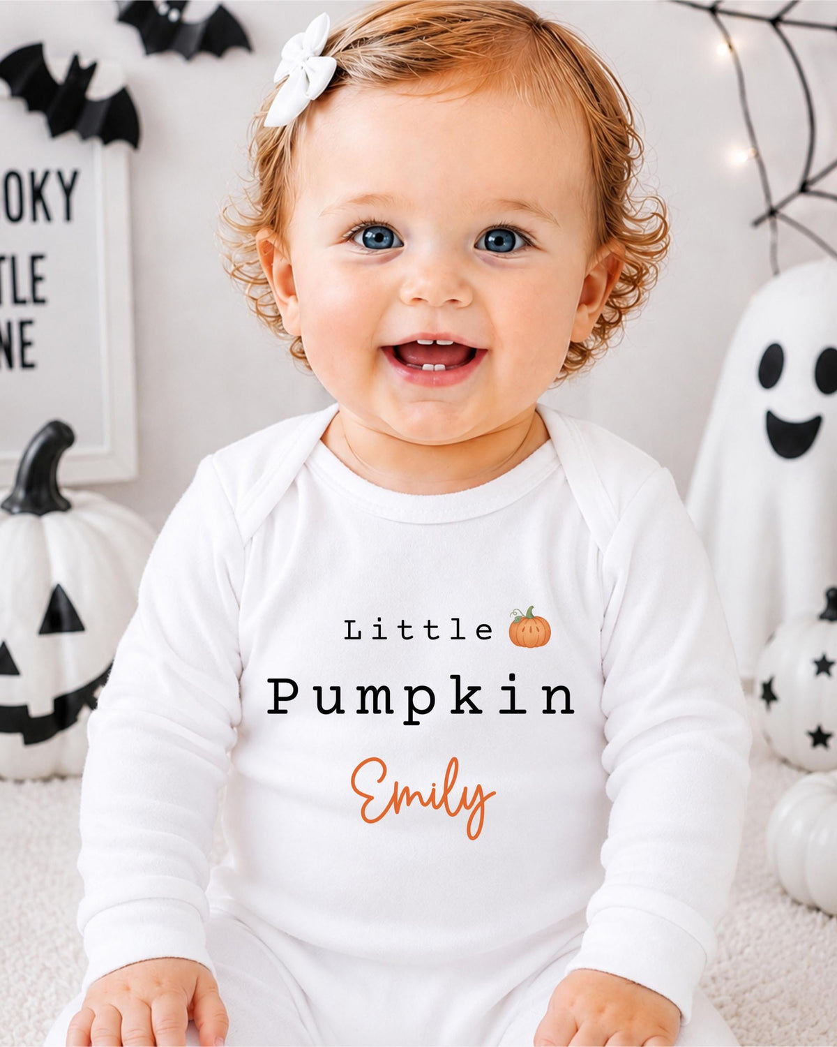 A smiling baby girl wearing a white onesie with the text "Little Pumpkin Emily" is sitting in front of Halloween decorations.