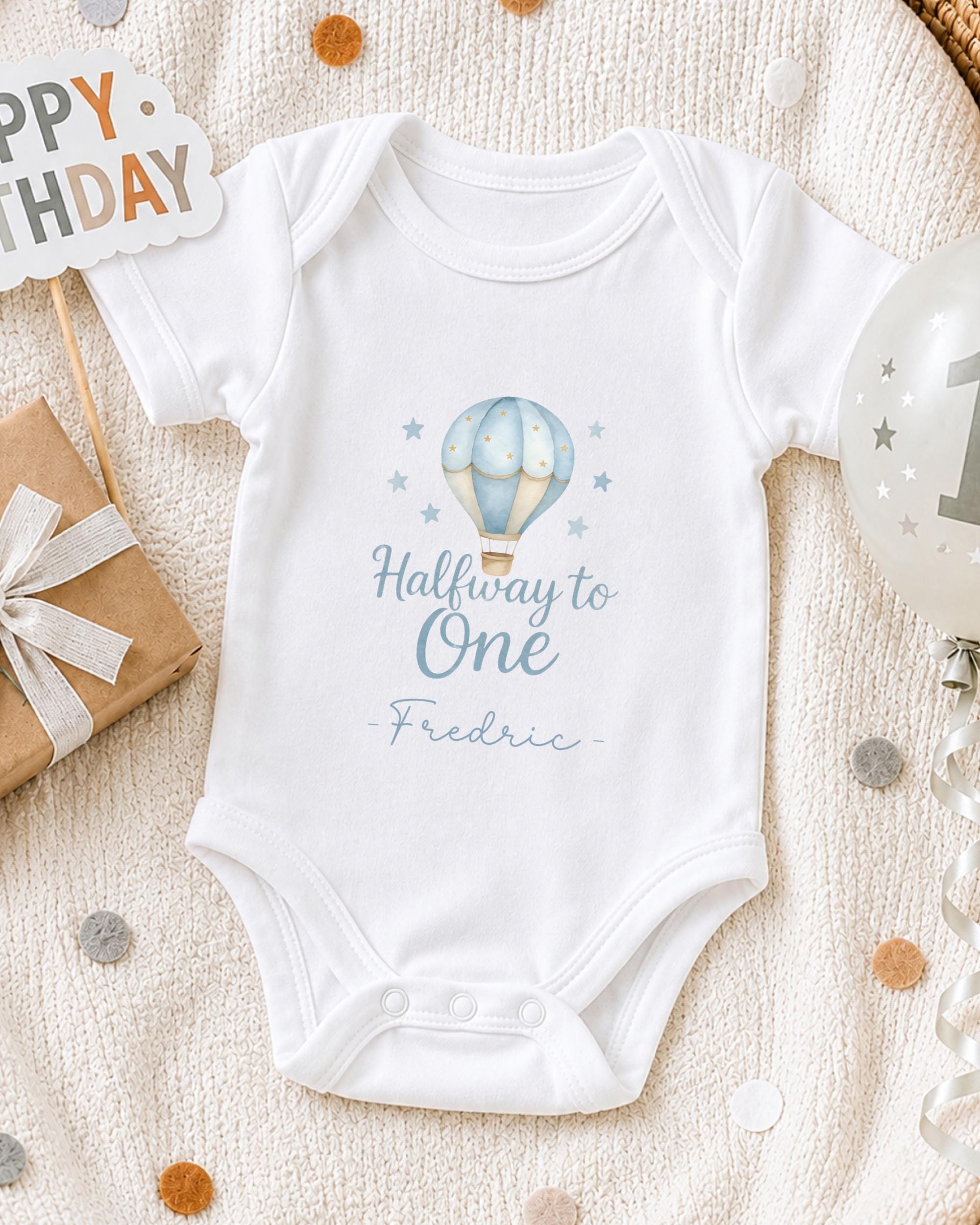 A white onesie with a blue hot air balloon design and the text "Halfway to One" printed on it, along with a "Fredric" signature, is displayed on a bed with a "Happy Birthday" sign and confetti.