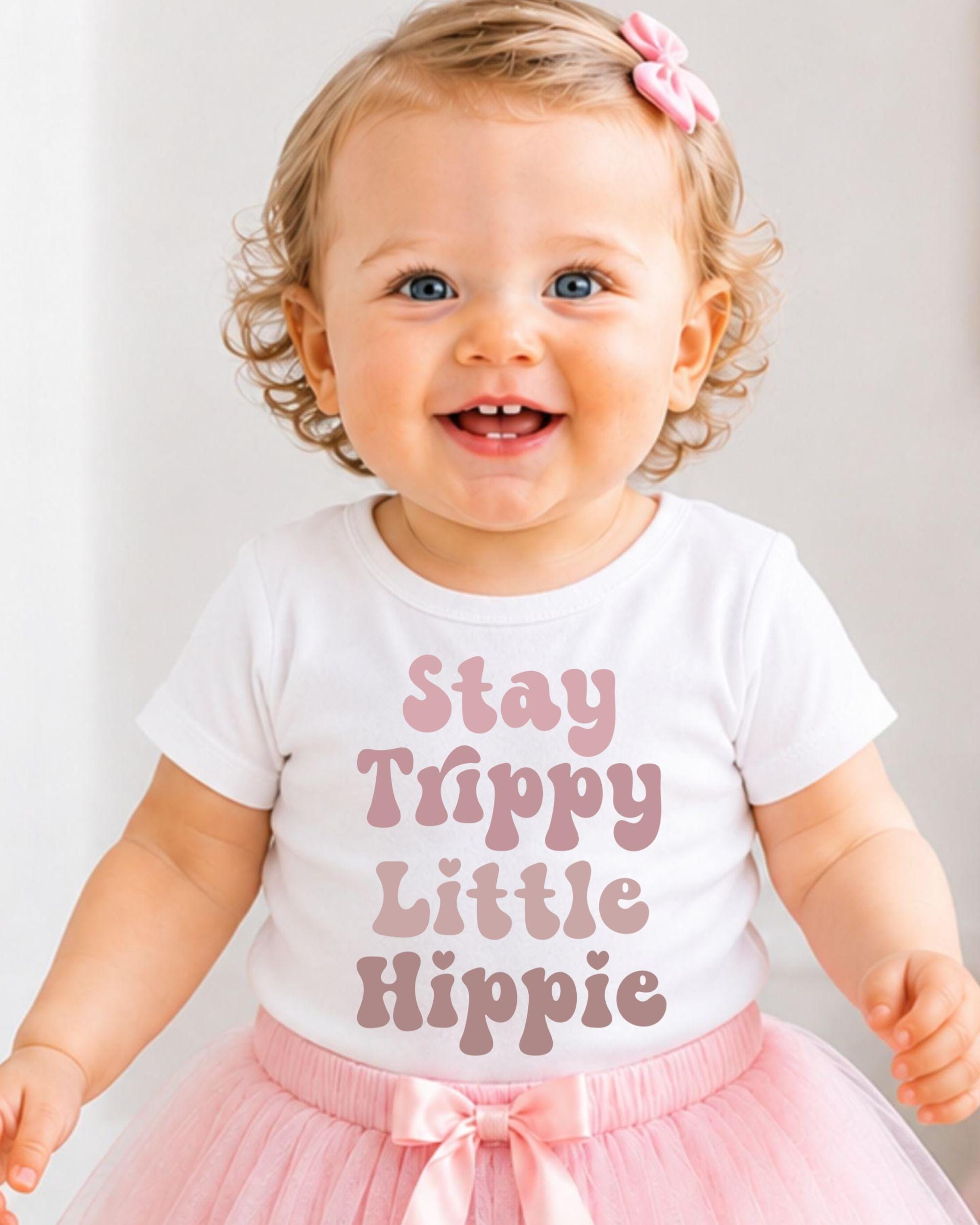 A smiling baby girl wearing a white t-shirt with the text "Stay Trippy Little Hippie" and a pink tutu skirt.