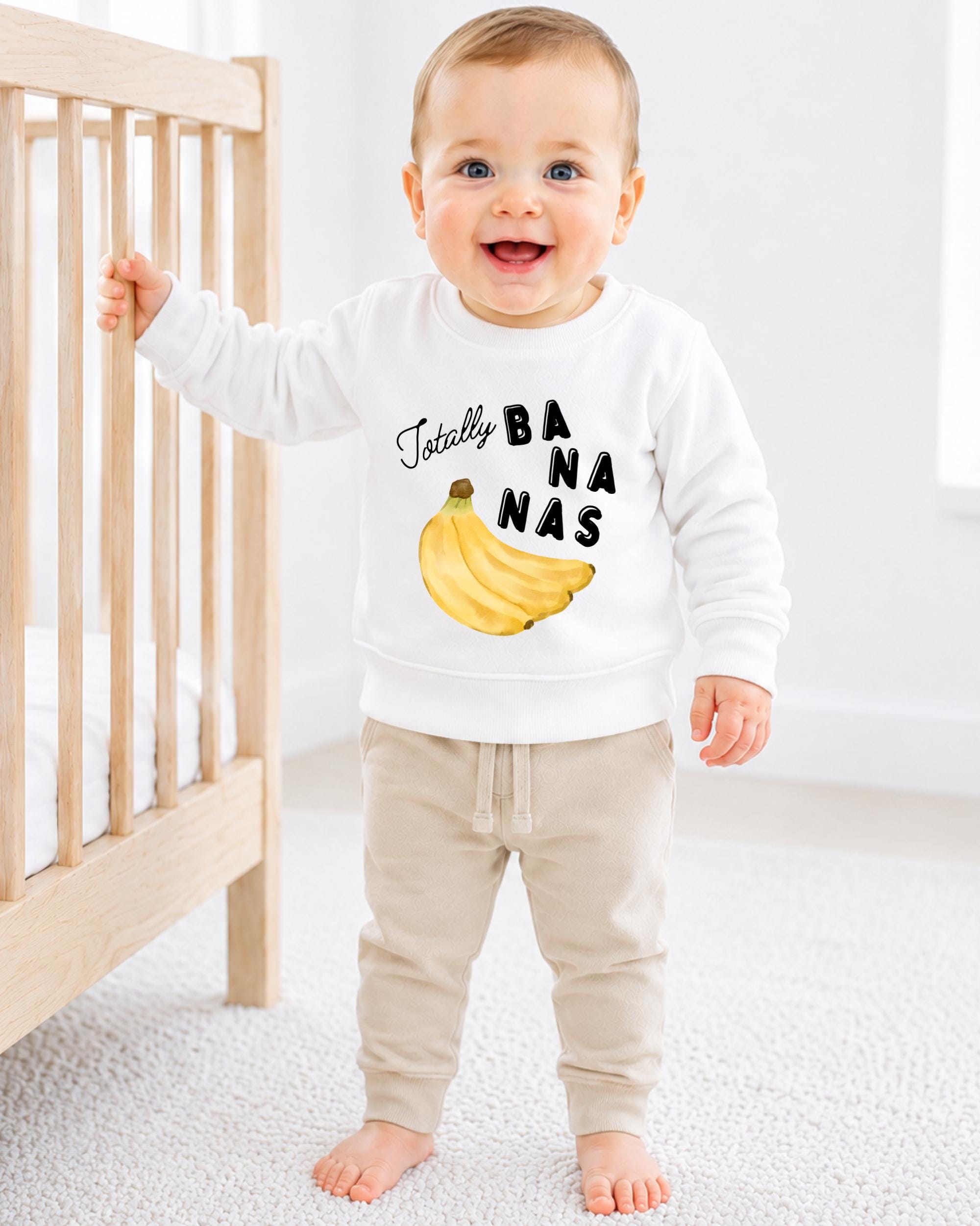 A smiling baby boy wearing a white sweatshirt with a banana graphic and the text "Totally BANANA" is standing next to a wooden crib.