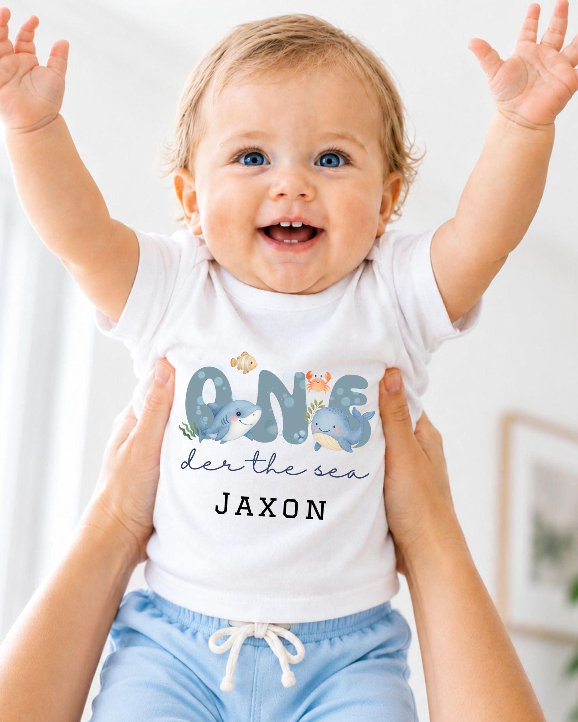 A smiling baby wearing a white onesie with a design of a shark and the text "One der the sea JAXON" is being held up by someone's arms.