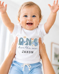 A smiling baby wearing a white onesie with a design of a shark and the text "One der the sea JAXON" is being held up by someone's arms.