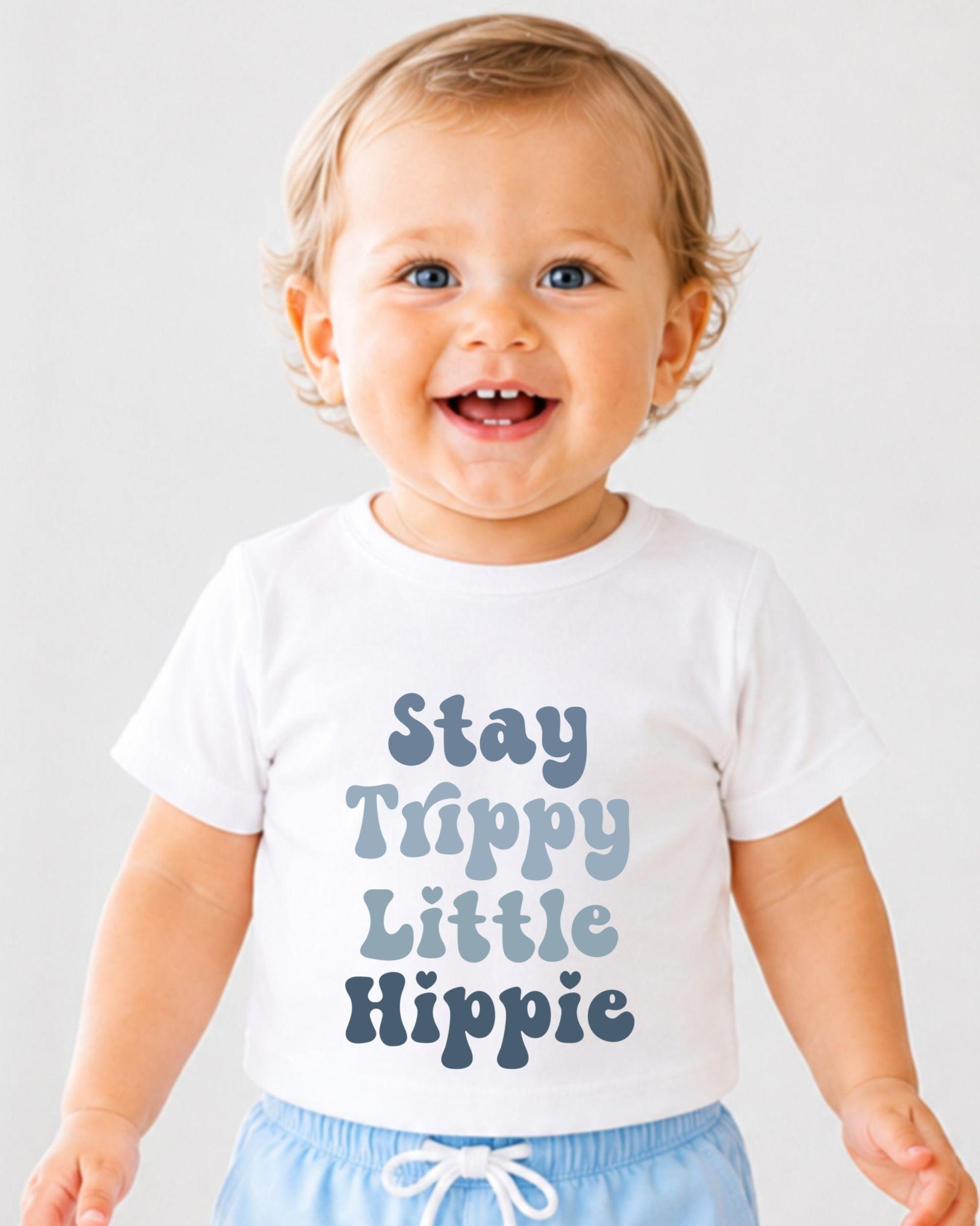 A smiling baby wearing a white t-shirt with the text "Stay Trippy Little Hippie" printed on it.