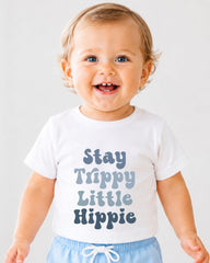 A smiling baby wearing a white t-shirt with the text "Stay Trippy Little Hippie" printed on it.