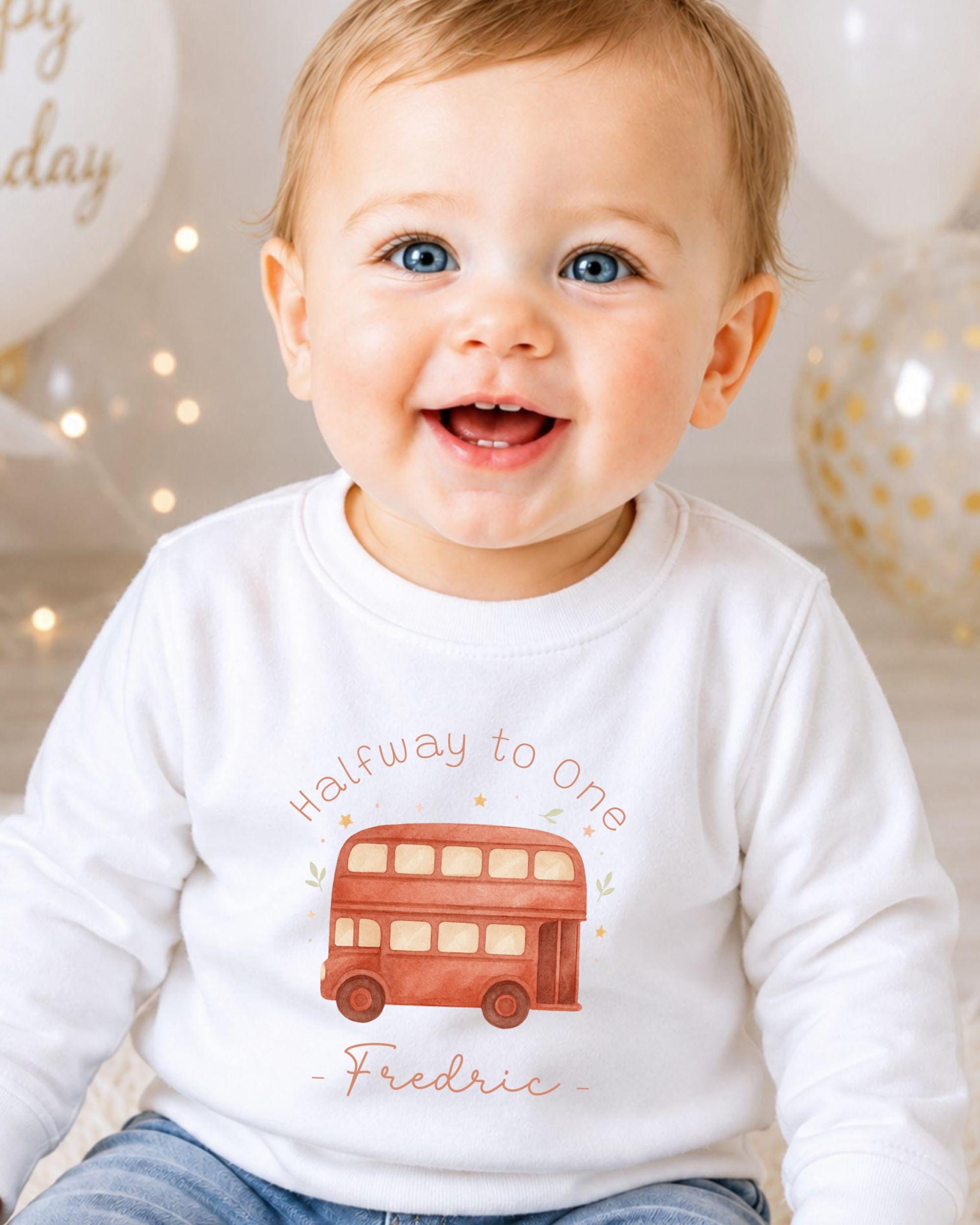 A smiling baby boy wearing a white shirt with a red double-decker bus graphic and the text "Halfway to one - Frederic" printed on it.