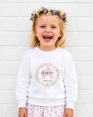 A young girl wearing a white sweatshirt with a floral wreath and the name "Aimee" printed on it, smiling and standing in front of a white brick wall.