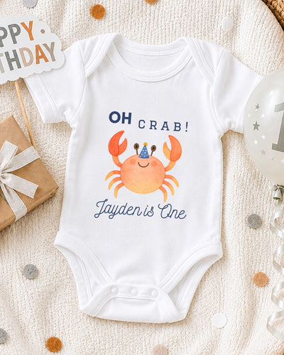 A white bodysuit with a crab design and the text "OH Crab! Jayden is One" printed on it, along with a birthday sign and a balloon.