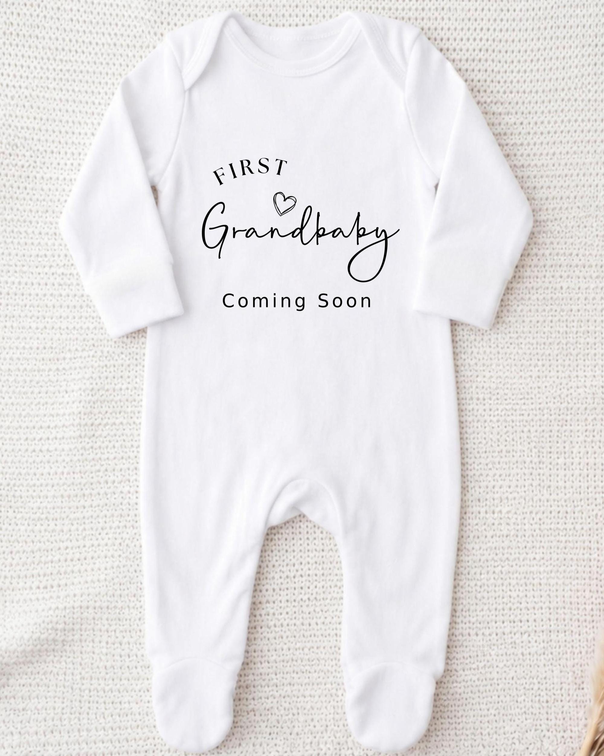 A white onesie with the text "FIRST Grandbaby Coming Soon" printed on it.