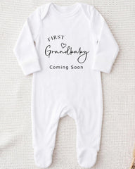 A white onesie with the text "FIRST Grandbaby Coming Soon" printed on it.