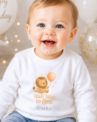 A smiling baby wearing a white shirt with a cartoon lion and the text "Half Way to One" and "SIMBA" printed on it.