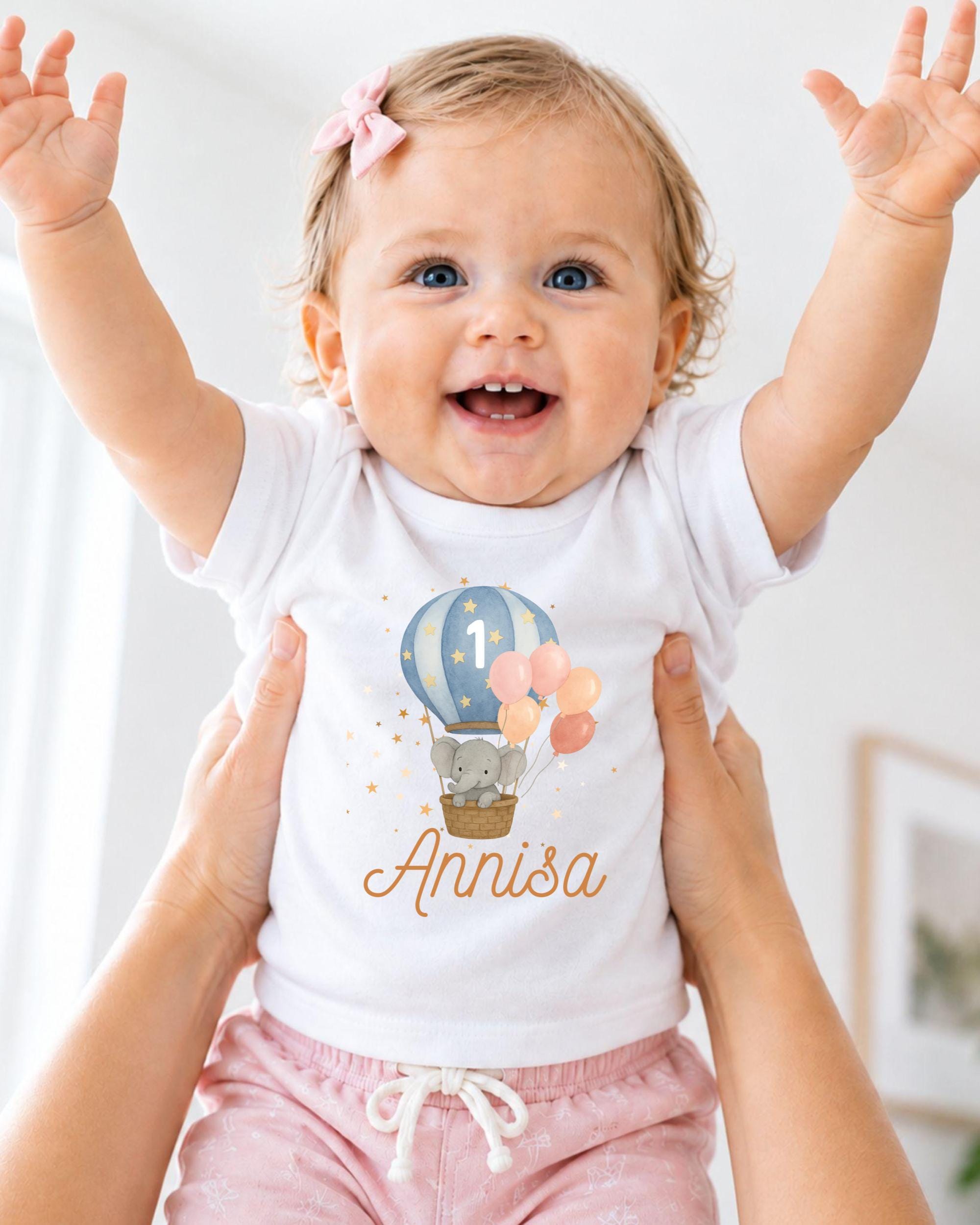 A smiling baby girl wearing a white t-shirt with a hot air balloon and elephant design, and pink shorts, with her arms raised in a joyful gesture.