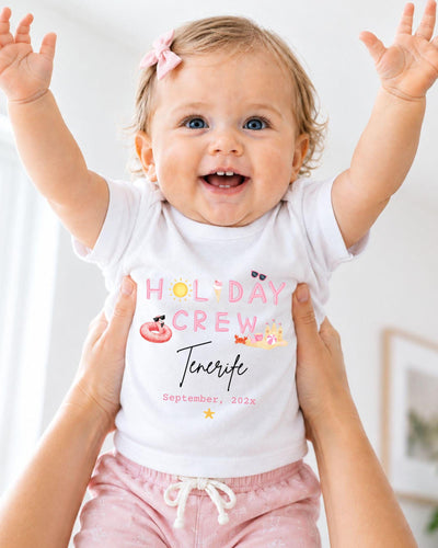 A smiling baby girl wearing a white t-shirt with the text "HOLIDAY CREW" and "Tenerife" printed on it, being held up by someone's arms.