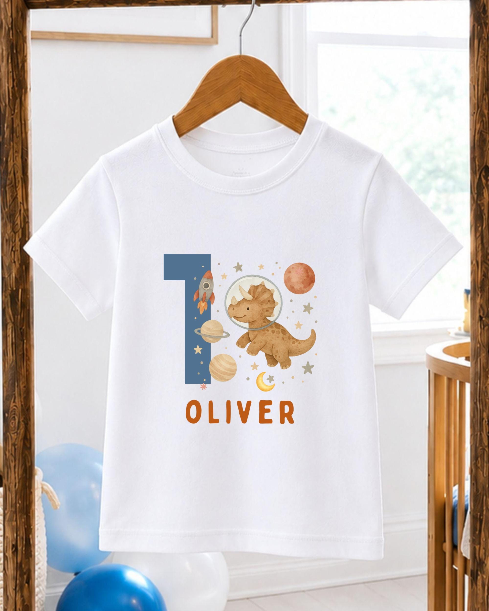 A white t-shirt with a dinosaur and space-themed design, along with the name "OLIVER" printed on it.