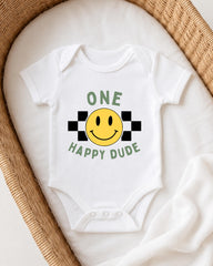 A white onesie with a yellow smiley face and the text "ONE HAPPY DUDE" printed on it, laid out on a white blanket.