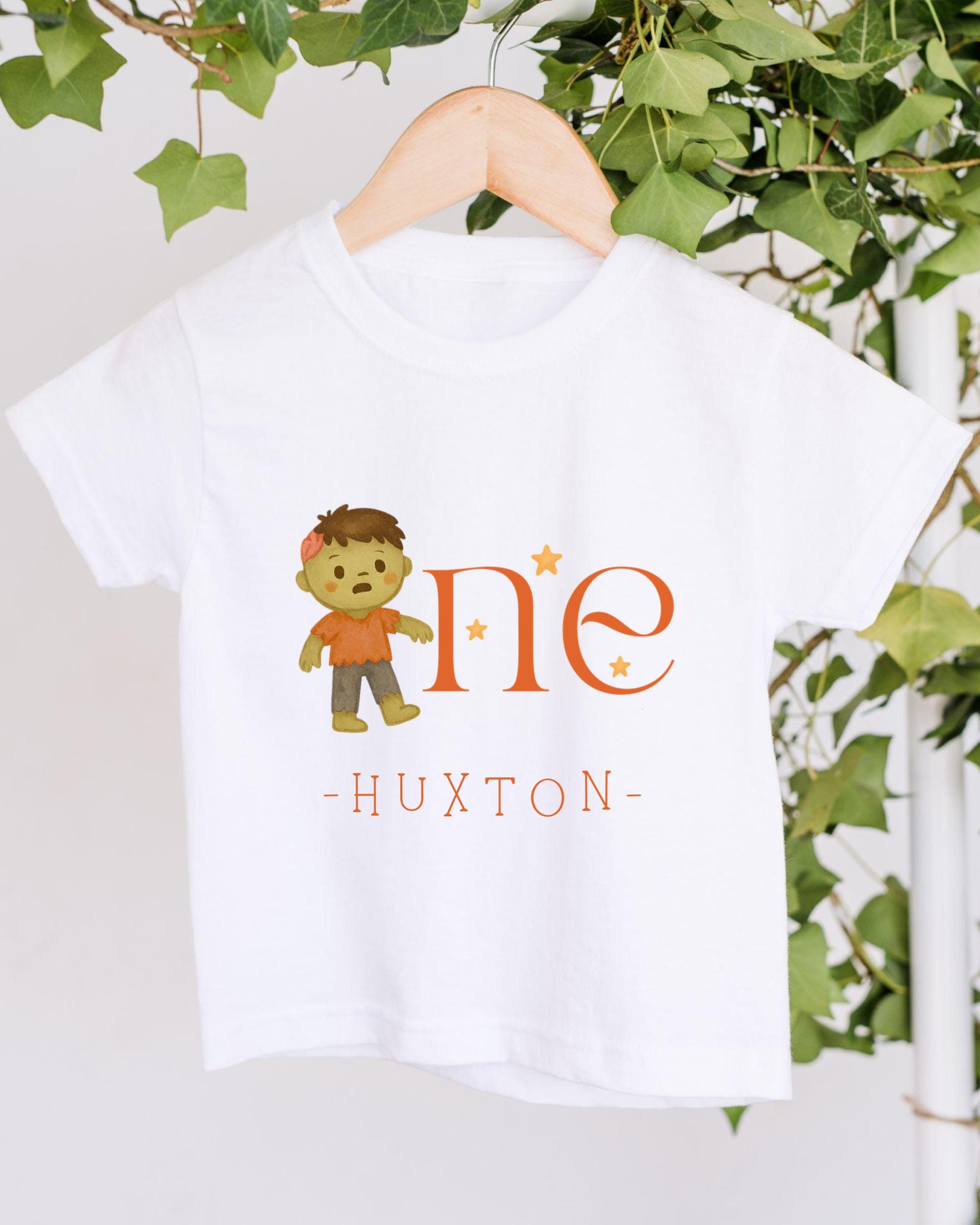 A white t-shirt with a cartoon character and the text "HUXTON" printed on it, hanging on a wooden hanger.